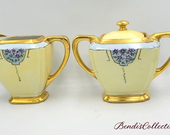 May include: A vintage cream-colored porcelain creamer and sugar set with gold trim and floral designs. The set features a delicate floral pattern in shades of blue and purple.