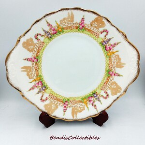 May include: White ceramic plate with a scalloped edge and gold trim. The plate features a decorative border with floral and architectural motifs in pink, green, and brown. The plate is displayed on a wooden stand. The text "BendisCollectibles" is at the bottom.