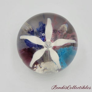May include: A clear glass paperweight with a white flower inside. The flower has five petals and is surrounded by blue, pink, and white swirls.
