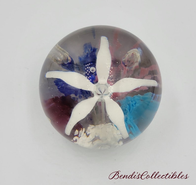 May include: A clear glass paperweight with a white flower inside. The flower has five petals and is surrounded by blue, pink, and white swirls.