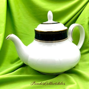 May include: White teapot with a black and gold trim. The teapot has a lid and a handle. The teapot is sitting on a green surface.