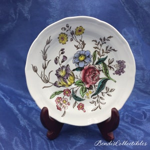 May include: White saucer with a floral design featuring pink, yellow, purple, and blue flowers. The flowers are surrounded by green leaves and brown stems.
