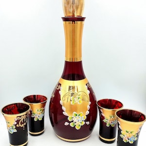 May include: A deep red glass decanter with a gold stopper and four matching glasses. The set features gold painted panels decorated with hand-painted floral motifs in white, blue, and green. The text BendisCollectibles is visible at the bottom.