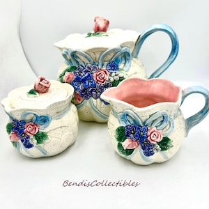 Puede incluir: Un juego de té de cerámica blanca con tetera, jarra de crema y azucarero. El juego está decorado con diseños florales azules y rosas y encaje blanco.