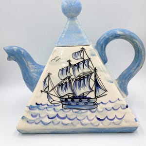 May include: A ceramic teapot with a triangular body and a light blue handle, spout, and lid. The teapot features a hand-painted design of a sailing ship on the ocean in shades of blue and white. The text "BendisCollectibles" is at the bottom.