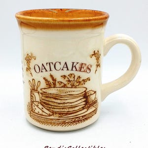 May include: Cream-colored ceramic mug with a brown rim and handle. The mug features the word "OATCAKES" above a brown illustration of oatcakes and other food items. The mug has a vintage aesthetic.