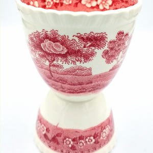 May include: A white and red ceramic egg cup with a landscape design. The egg cup features a scalloped rim and a pedestal base. The interior and base have floral patterns. The design is in a classic toile style.