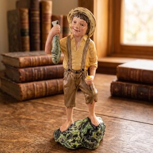 Puede incluir: Una figura de cerámica de un joven descalzo con sombrero de paja, camisa amarilla y pantalones marrones con tirantes, de pie sobre una roca verde y negra. La figura sostiene un pez verde en la mano derecha. El texto Bendi'sCollectibles es visible.