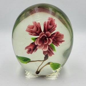 May include: A clear glass paperweight with a pink flower inside. The flower has green leaves and a stem.