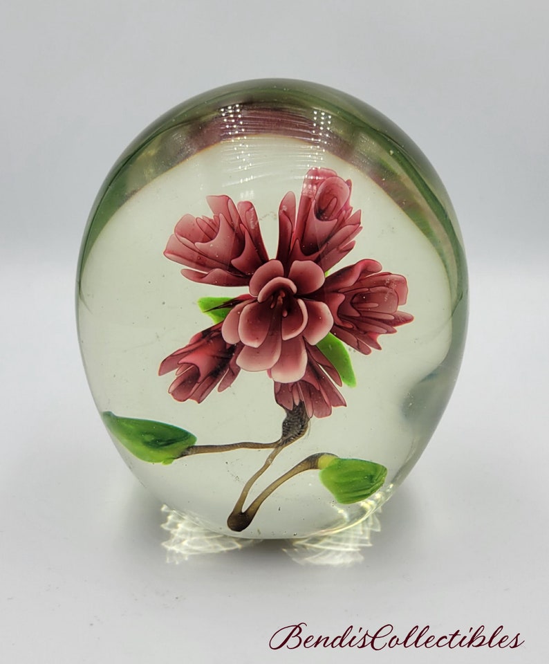 May include: A clear glass paperweight with a pink flower inside. The flower has green leaves and a stem.