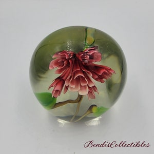 May include: A clear glass paperweight with a pink flower and green leaves inside. The flower is in bloom and has a delicate, intricate design.