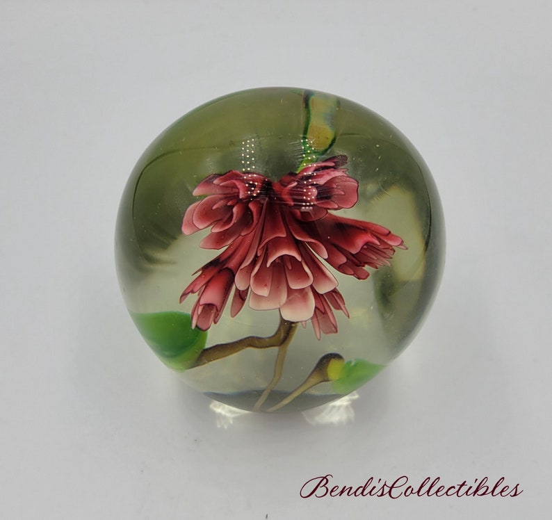 May include: A clear glass paperweight with a pink flower and green leaves inside. The flower is in bloom and has a delicate, intricate design.