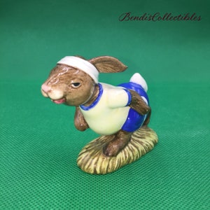 Puede incluir: Una figurita de conejo marrón y blanco con un uniforme deportivo azul y blanco, corriendo sobre una base de hierba marrón. La figurita forma parte de una colección de personajes de Beatrix Potter.