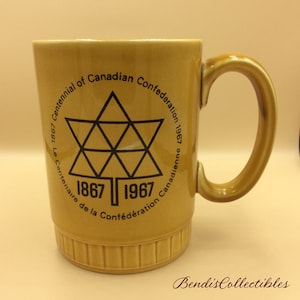 Puede incluir: Una taza de cerámica amarilla con un gráfico negro que conmemora el centenario de la Confederación Canadiense de 1967. El gráfico presenta una hoja de arce estilizada con los años 1867 y 1967.
