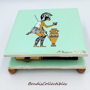 Puede incluir: Una caja de música cuadrada vintage con una tapa verde claro que presenta un diseño de figura griega clásica. La figura está representada en negro, blanco y dorado, sosteniendo una copa. La caja descansa sobre pequeños pies de madera.