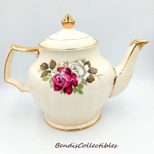 May include: A cream-coloured vintage teapot with a floral design featuring red and white roses, green leaves, and brown accents. The teapot has gold trim on the handle, spout, and lid. The base of the teapot is also trimmed in gold.