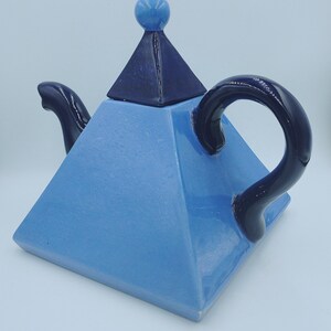 May include: A light blue ceramic teapot with a pyramid-shaped body and lid. The lid is dark blue and topped with a small blue sphere. The handle and spout are also dark blue. The text "BendisCollectibles" is at the bottom.