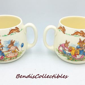 May include: Two cream-colored ceramic mugs with a whimsical bunny design. The mugs feature illustrations of bunnies playing, with a running bunny border. The mugs have a curved shape and a handle. The text "BendisCollectibles" is at the bottom.