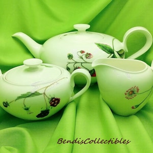 Puede incluir: Un juego de té de cerámica blanca con tetera, azucarero y jarrita para la leche. Cada pieza está decorada con un diseño floral y de bayas en tonos rojos, verdes y marrones. El juego se exhibe sobre un fondo de tela verde.