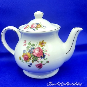 Może przedstawiać: Biały czajnik z kwiatowym wzorem z różowymi, żółtymi i niebieskimi kwiatami. Czajnik ma złotą obwódkę wokół pokrywki i dzióbka.