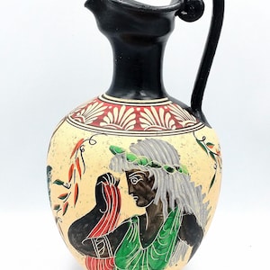 May include: A ceramic pitcher featuring a black neck and handle, with a cream-colored body decorated with a red and white floral pattern at the top and a central illustration of a figure wearing a green garment and a green wreath on their head.