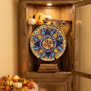 May include: A round decorative ceramic plate featuring a vibrant, multi-colored mandala pattern with intricate floral and geometric motifs in shades of blue, red, yellow, and green, displayed on a stand atop a stack of three books inside a cabinet.