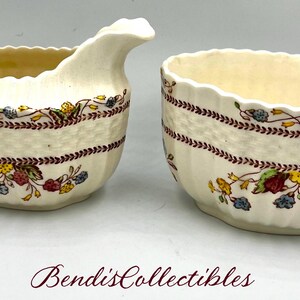 May include: A vintage cream-coloured creamer and sugar bowl set with a floral pattern in shades of red, yellow, and blue. The set features a delicate, woven design and a red and brown trim.