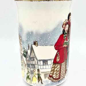 May include: A white ceramic mug with a gold rim, featuring a winter scene illustration. The artwork depicts figures in period clothing, snow-covered buildings, and a snowy sky. The mug has the text "BendisCollectibles" at the bottom.