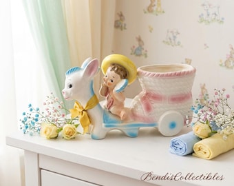 May include: A ceramic planter shaped like a donkey pulling a woven-textured boot cart. The donkey is white with blue accents and a yellow ribbon. A small figure wearing a yellow hat sits on the donkey, and the boot cart features pink ribbon details.