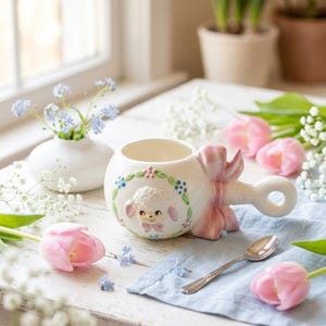 Könnte beinhalten: Ein cremefarbener Keramikbecher mit einem Relief eines Lammgesichts, das von einem Blumenkranz umgeben ist. Der Becher hat einen ringförmigen Henkel mit einer großen, modellierten rosa Schleife an der Seite. Ein kleiner silberfarbener Metalllöffel liegt daneben.