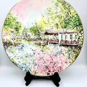 Op de afbeelding: Decoratieve plaat met een serene Aziatische landschap met een paviljoen, bomen en water. De plaat heeft een gouden rand en een roze en witte lucht. De scène is geschilderd in een zachte aquarel stijl. De plaat wordt gepresenteerd op een zwarte standaard.
