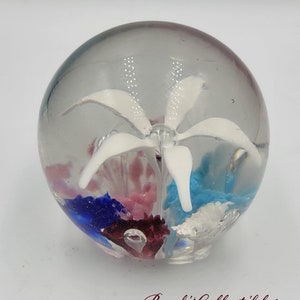 May include: A clear glass paperweight with a white flower inside. The flower is surrounded by blue, pink, and red glass pieces.