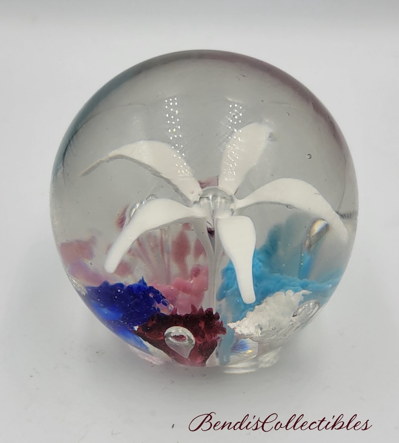 May include: A clear glass paperweight with a white flower inside. The flower is surrounded by blue, pink, and red glass pieces.