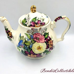 Może przedstawiać: Biały czajnik z kwiatowym wzorem. Czajnik ma złotą obwódkę i pokrywkę z kwiatowym wzorem. Czajnik jest ozdobiony różnymi kwiatami, w tym różami, liliami i stokrotkami.