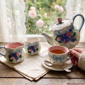 May include: A ceramic tea set featuring a teapot, creamer, sugar bowl, and teacup with saucer. The pieces are cream-colored with embossed blue ribbons, pink rose accents, and purple floral clusters. The teacup interior is pink, and a silver spoon rests on the saucer.