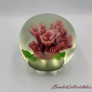 May include: A glass paperweight with a pink flower inside. The flower is surrounded by green leaves. The paperweight is clear and has a shiny finish.