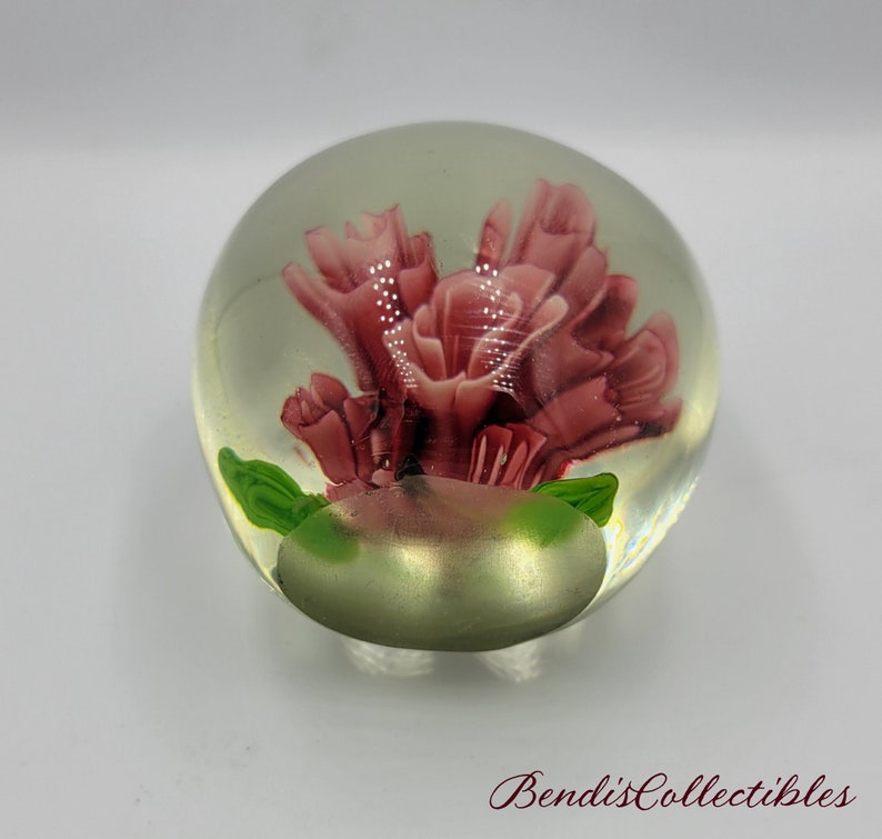 May include: A glass paperweight with a pink flower inside. The flower is surrounded by green leaves. The paperweight is clear and has a shiny finish.