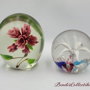 May include: Two glass paperweights with floral designs. One paperweight features a pink flower with green leaves, while the other features a white flower with blue and pink accents.