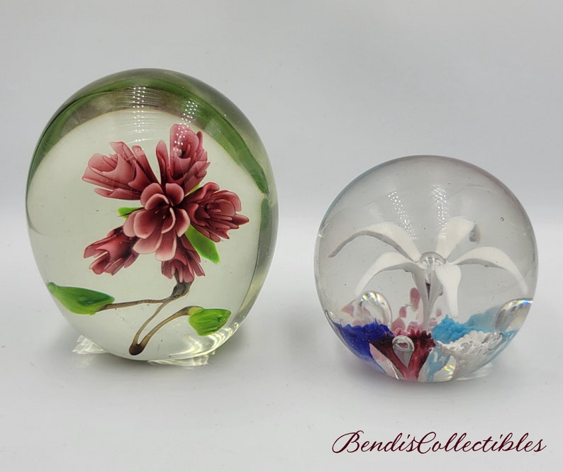 May include: Two glass paperweights with floral designs. One paperweight features a pink flower with green leaves, while the other features a white flower with blue and pink accents.