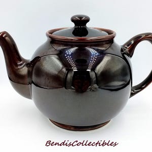 May include: A round, dark brown glazed ceramic teapot with a matching lid, a curved spout, and a sturdy handle. The surface has a high-gloss finish with subtle reddish-brown undertones. The text BendisCollectibles is printed in dark brown at the bottom.