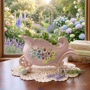 May include: A light pink ceramic wheelbarrow planter with a textured surface, gold trim, and a floral relief design. It contains green succulents and sits on a cream-coloured crocheted doily with small speckled eggs and two silver bird figurines.