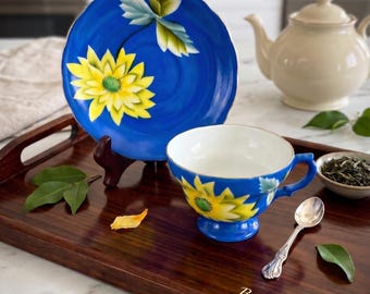 Puede incluir: Un juego de taza y platillo azul vibrante con girasoles amarillos pintados a mano y hojas verdes. El conjunto se exhibe en una bandeja de madera oscura con el texto Bend'sCollectibles grabado en la esquina, junto a una cuchara plateada.