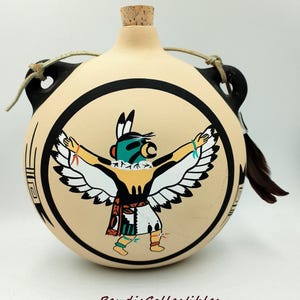 May include: A tan-colored, round ceramic flask with a cork stopper and a black rim. The flask features a Native American design of a figure with outstretched arms and white wings. The figure is wearing a turquoise mask and a colorful outfit. The text "BendisCollectibles" is at the bottom.