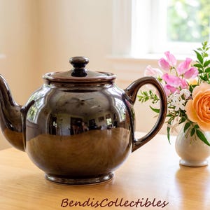 May include: A glossy dark brown ceramic teapot with a rounded body, a curved handle, a spout, and a matching lid. The surface of the teapot is highly reflective, showing a distorted reflection of the room. The text BendisCollectibles is visible.