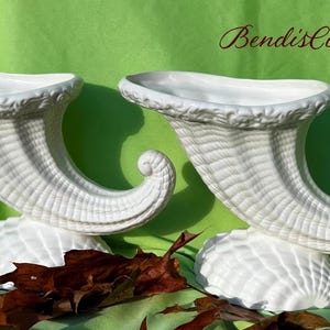 May include: Two white ceramic cornucopia vases. Each vase has a curved horn shape with a textured surface and sits on a seashell-shaped base. The background is green with the text "BendisCollectibles".