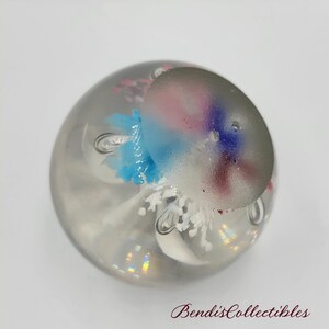 May include: A clear glass paperweight with a white, blue, pink, and purple flower design inside. The flower design is reminiscent of a jellyfish.