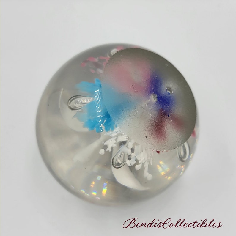 May include: A clear glass paperweight with a white, blue, pink, and purple flower design inside. The flower design is reminiscent of a jellyfish.
