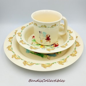 Op de afbeelding: Een crèmekleurige kinderservies set met een mok, een kom en een bord. De set is versierd met een herhalend patroon van gele konijnen en een centrale illustratie van stripfiguren. De tekst "BendisCollectibles" staat onderaan.