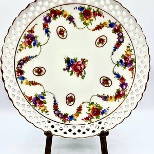 Puede incluir: Un plato de porcelana blanca con borde calado y borde dorado. El plato está decorado con coloridos motivos florales en rojo, morado, azul y amarillo. El plato se exhibe sobre un soporte de bronce. El texto "BendisCollectibles" está en la parte inferior.