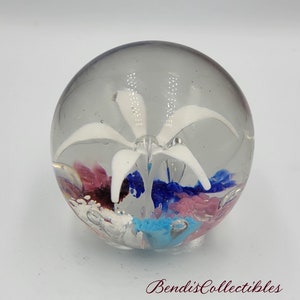 May include: A clear glass paperweight with a white flower inside. The flower has five petals and is surrounded by swirls of pink, blue, and turquoise glass.